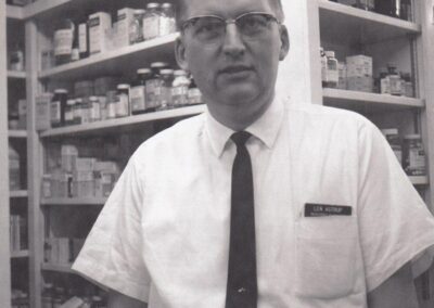 A photo of Len Astrup in a white button up shirt, and black tie, standing in front of shelves of medication.