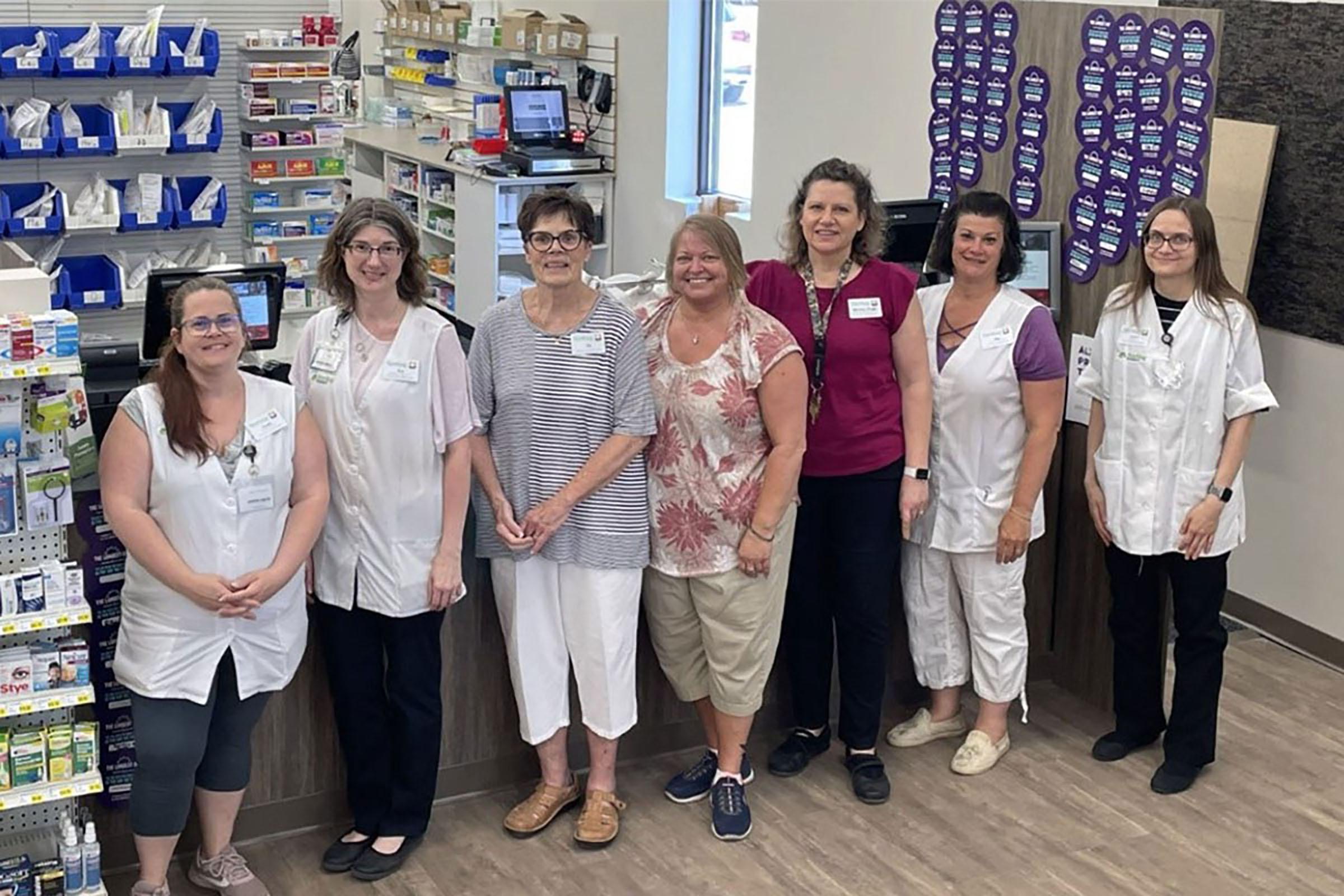 Pharmacy employees standing in front of their counter, and shelves of medication.